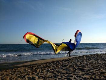 Man carrying parachute while walking on shore at beach against sky