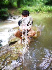 Young man splashing water while crouching on rock at forest