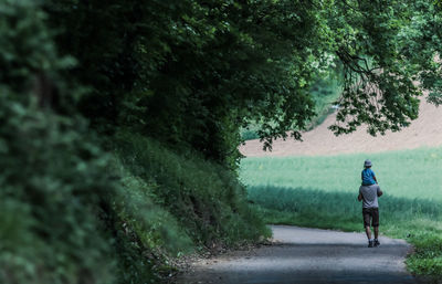 Rear view of father carrying daughter while walking on footpath by trees