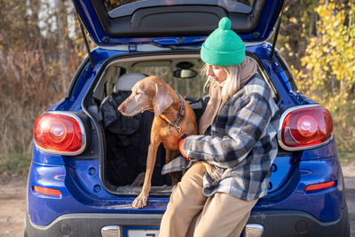 Pet owner woman in road trip holding dog on leash sitting in car trunk on nature.