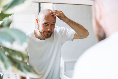 Young adult bearded man looking in mirror in bathroom touching head worried about about hair loss