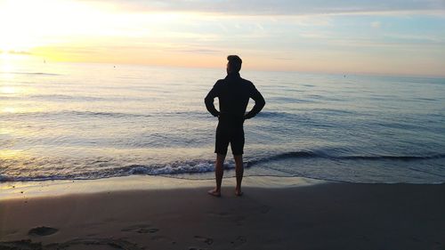 Rear view of man with hands on hips standing on shore during sunset