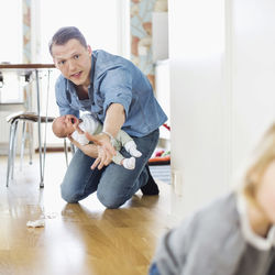 Father holding crying baby while stopping daughter on floor at home