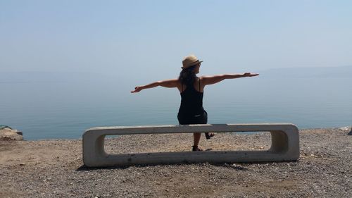 Rear view of woman sitting on concrete seat by lakeshore against sky