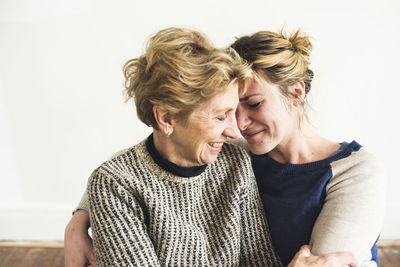 Close-up of smiling mother and daughter sitting against wall at home