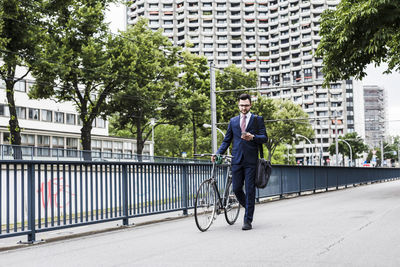 Businessman with bicycle walking in the city, using smart phone