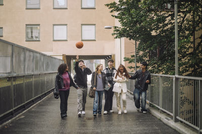 Boy playing basketball while walking with male and female friends on footpath