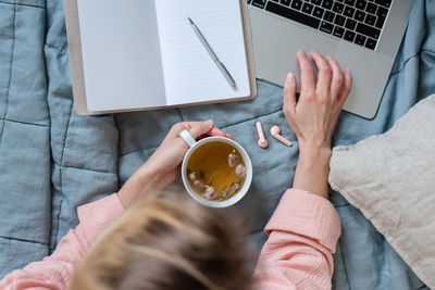 Overhead scene in pastel colors of a woman using a laptop, making notes, and drinking tea.