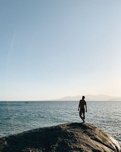Rear view of shirtless man standing on rock by sea against clear sky