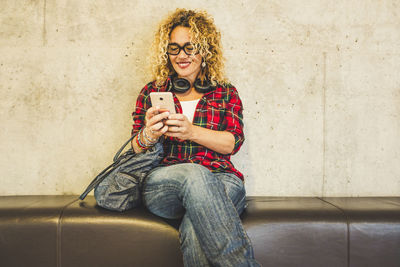 Smiling woman holding smart phone while sitting against wall