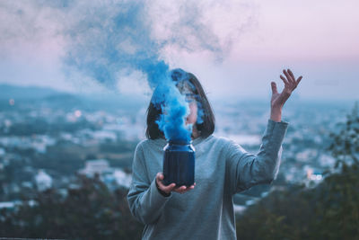 Woman holding container with blue smoke against sky