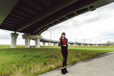 Full length of young woman standing on footpath by field under bridge