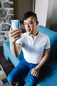 High angle of teenage latin boy with down syndrome taking self shot on smartphone while sitting on sofa at home