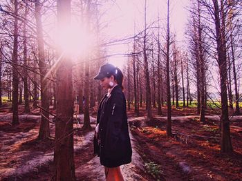 Side view of young woman standing amidst bare trees in forest on sunny day