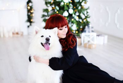Young woman embracing dog against christmas tree at home