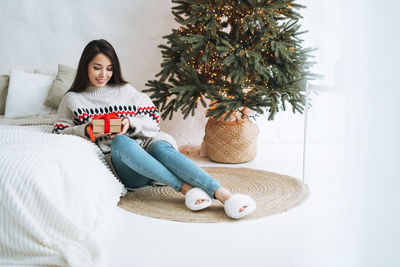 Young asian woman with long hair in cozy sweater with gift box in bedroom with christmas tree 