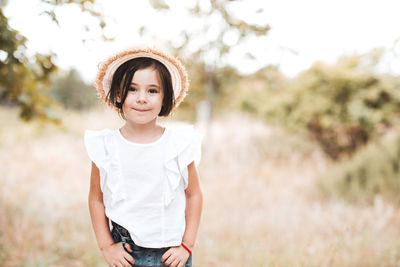 Smiling child girl 4-5 year old wearing straw hat, white top and denim shorts posing in meadow