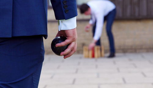 Midsection of man holding ball against friend arranging bowling pins on footpath