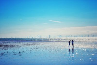 Full length of boys standing on shore at beach against blue sky during sunny day