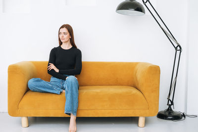 Young thinking woman girl with dark long hair in jeans on yellow sofa in minimalistic interior