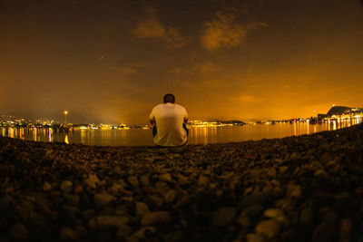 Rear view of man sitting at lakeshore during sunset