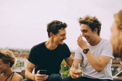 Happy man showing mobile phone to friend having drink at terrace during party