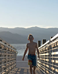 Full length portrait of shirtless boy walking on footbridge at beach against sky