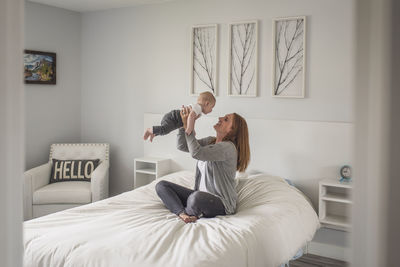 Side view of happy mother picking up son while sitting on bed against wall at home