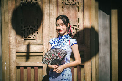Portrait of smiling teenager girl holding hand fan against wooden door