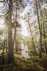 Mid adult bride holding bouquet looking at groom standing amidst trees in forest