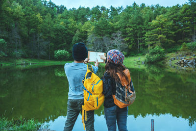 Rear view of couple holding book while standing by lake at forest