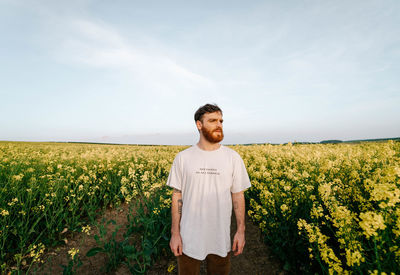 Young man looking away while standing amidst flowers against sky