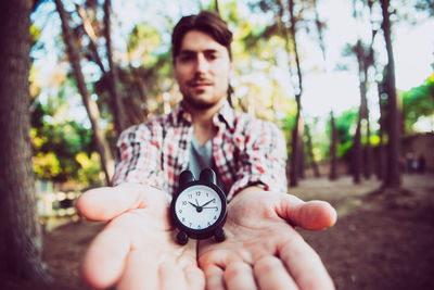 Portrait of young man holding clock against trees