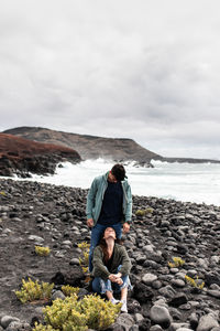 Full body of unrecognizable young female traveler sitting on rocky beach of powerful ocean and looking up on standing boyfriend during vacation in lanzarote