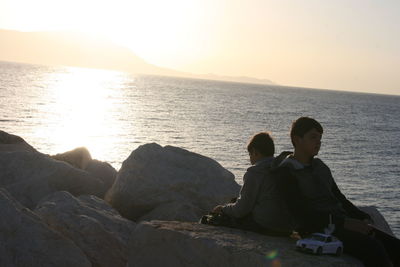 People sitting on rock by sea against sky during sunset