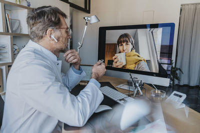 Mature doctor talking to patient on video call over computer while sitting at doctor's office