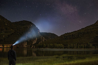 Rear view of man with illuminated headlamp on lakeshore against star field