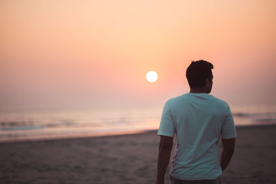 Rear view of man standing at beach against sky during sunset