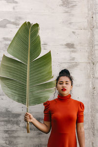 Young woman in turtleneck red dress holding green banana leaf in front of wall