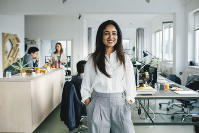 Portrait of confident smiling businesswoman standing with hands in pockets at office