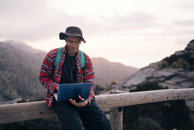 Young man using laptop while sitting on railing at sunset