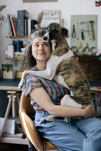 Smiling woman embracing dog while sitting on chair at home