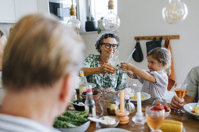 Grandmother toasting drinks with granddaughter sitting at dining table during dinner with family at home