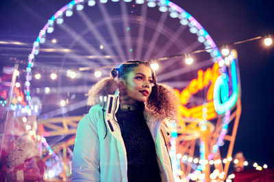 Young woman standing against illuminated carousel in amusement park