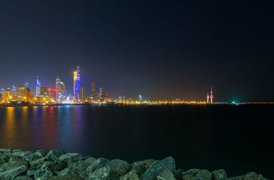 Illuminated buildings by sea against sky at night