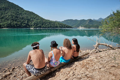 Back view of company of friendly people relaxing on tree log near lake and enjoying sunny day during vacation together