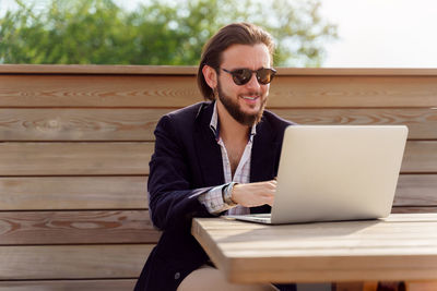 Smiling businessman using laptop while sitting on bench outdoors