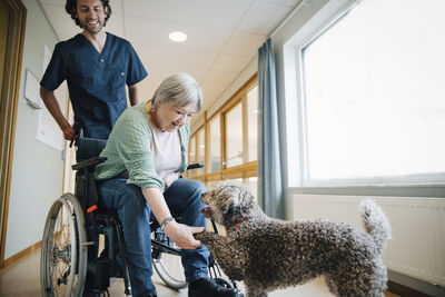 Smiling male nurse looking at disabled senior woman on wheelchair giving handshake to dog in alley