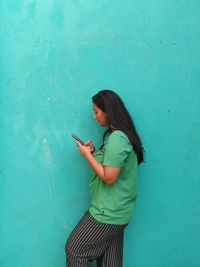 Young woman using mobile phone while standing against blue wall