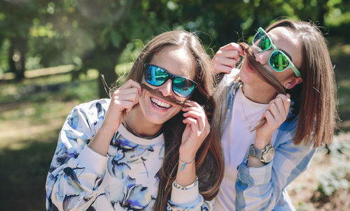 Portrait of friends wearing sunglasses while holding their hair as mustache at park